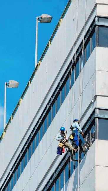 Black and white view from below of a steeplejack wearing a protective helmet and using professional gear to clean the glass wall of a modern tower
